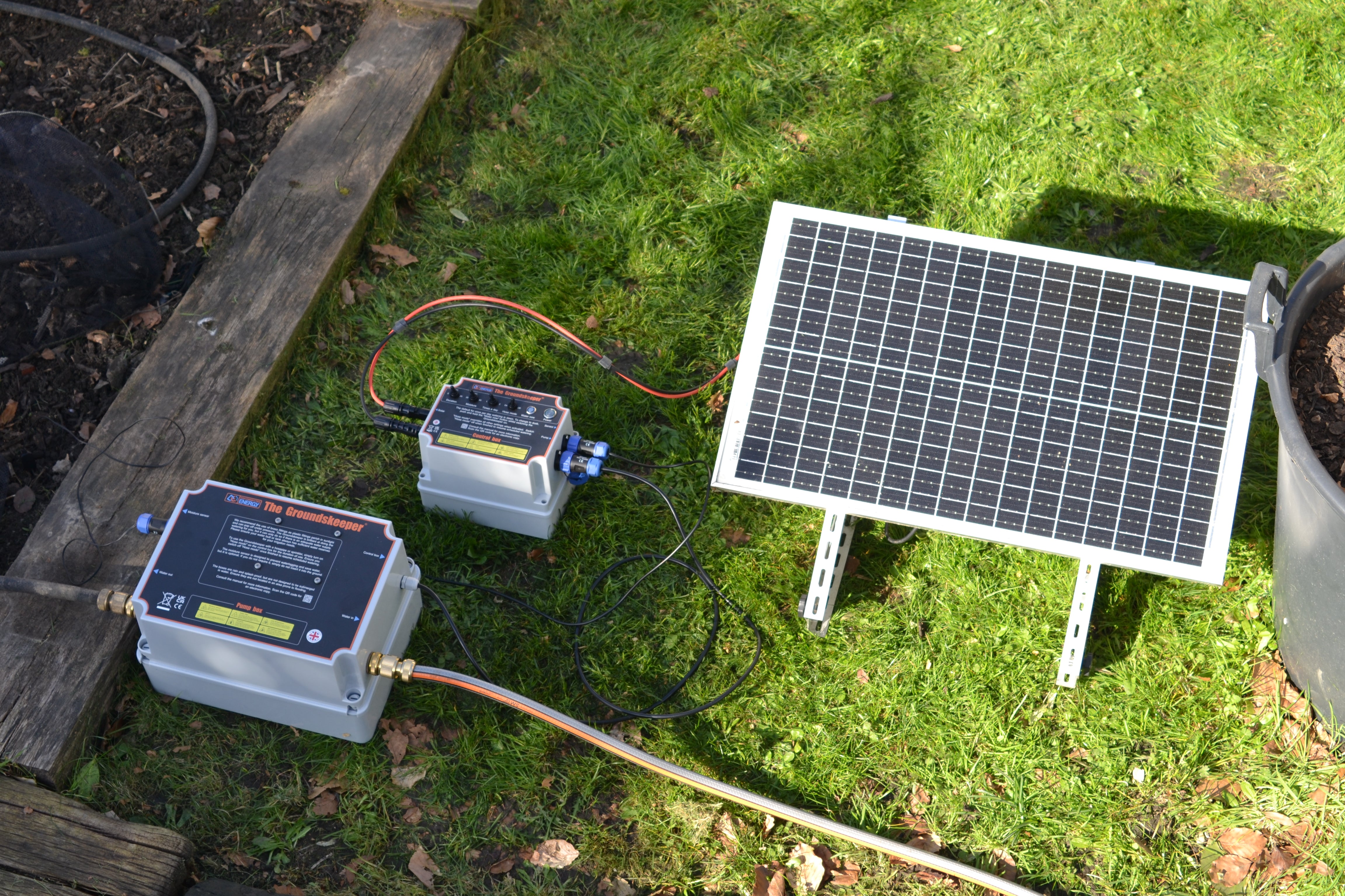 Groundskeeper garden water pump system comprising a solar panel, control box with switches on top and pump box connected to garden hose. All sitting on grass. 