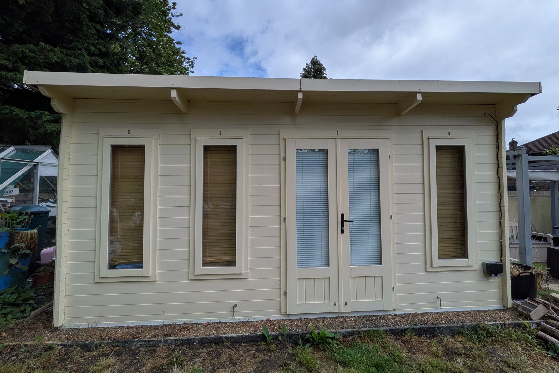 Small cream painted wooden cabin with windows, a door and an exterior power outlet, in a garden setting