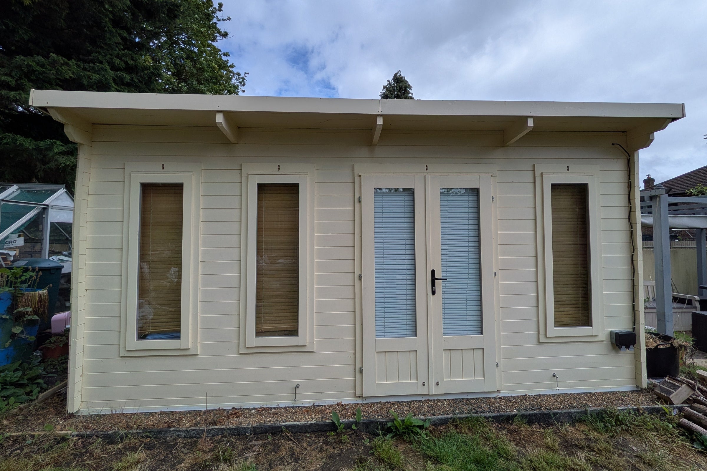 Small cream painted wooden cabin with windows, a door and an exterior power outlet, in a garden setting